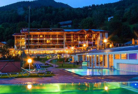 Ein hell erleuchtetes Hotel mit Holzbalkonen ist in der Abenddämmerung zu sehen, umgeben von Bäumen und Hügeln, mit Außenpools und Liegestühlen im Vordergund.