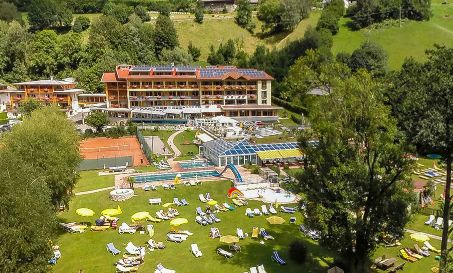 Ein großer Hotelkomplex mit Außenpools, Tennisplätzen und Liegestühlen ist von grünen Rasenflächen und Bäumen in einer hügeligen Gegend umgeben.