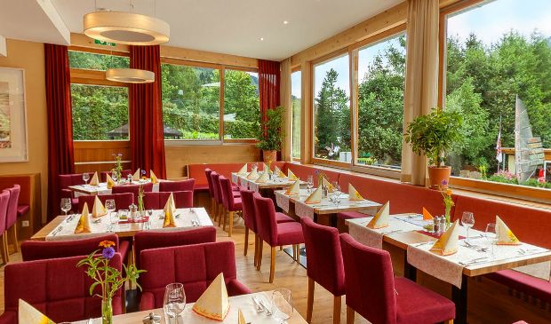 Ein heller Restaurantbereich mit roten Stühlen, gedeckten Tischen mit gefalteten Servietten, großen Fenstern und Topfpflanzen.