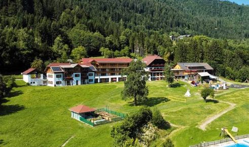 Eine Ansammlung von Gebäuden liegt auf einem grünen Hügel, umgeben von dichtem Wald und offenen Grasflächen, mit einem eingezäunten Bereich und kleinen Gebäuden in der Nähe.