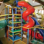 Indoor-Spielplatz mit einer großen roten Spiralrutsche, Klettergerüsten, Sicherheitsnetzen und verschiedenen Spielgeräten in einem hellen Raum.