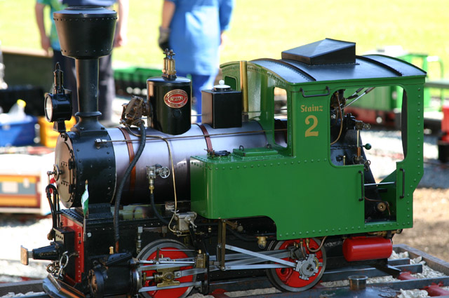 Eine kleine grün-schwarze Modell-Dampflokomotive mit der Aufschrift "Stainz 2" steht im Freien auf einem Gleis, und im unscharfen Hintergrund sind Menschen zu sehen.