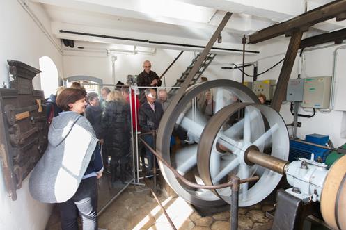 Eine Gruppe von Personen beobachtet ein großes sich drehendes Schwungrad in einem Maschinenraum mit industriellen Geräten und Schalttafeln.