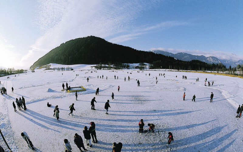 Eine weite Ansicht von Menschen, die auf einem großen zugefrorenen See Schlittschuh laufen und spazieren gehen, mit einem bewaldeten Hügel und Bergen im Hintergrund unter einem klaren Himmel.