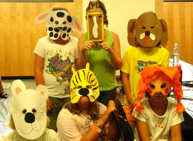 Sechs Kinder tragen handgefertigte Tiermasken und sitzen bzw. stehen gemeinsam im Raum und posieren für das Foto. Im Hintergrund sind Holzvertäfelungen und ein Tisch zu sehen.