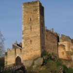 Eine steinerne Burgruine mit einem hohen rechteckigen Turm und einem gewölbten Eingang liegt auf einem felsigen Hügel, umgeben von Bäumen und teilweise eingestürzten Mauern unter einem klaren Himmel.