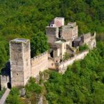 Luftaufnahme einer alten steinernen Burgruine, umgeben von dichtem grünen Wald, mit hohen Türmen und teilweise eingestürzten Mauern.