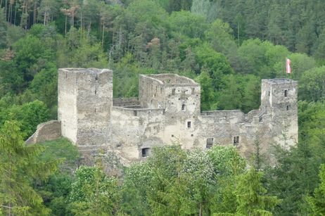 Eine steinerne Burgruine mit drei Haupttürmen steht inmitten eines dichten grünen Waldes; auf einem Turm ist eine österreichische Flagge zu sehen.