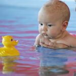 Zwei Erwachsene halten ein Baby in einem Schwimmbecken, während das Baby eine gelbe Gummiente ansieht, die auf dem Wasser schwimmt.