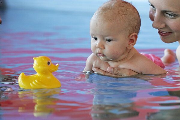 Zwei Erwachsene halten ein Baby in einem Schwimmbecken, während das Baby eine gelbe Gummiente ansieht, die auf dem Wasser schwimmt.