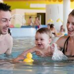 Ein Mann, eine Frau und ein Baby spielen mit einer gelben Gummiente in einem Schwimmbecken, umgeben von anderen Menschen.