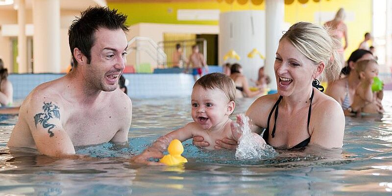 Ein Mann, eine Frau und ein Baby spielen mit einer gelben Gummiente in einem Schwimmbecken, umgeben von anderen Menschen.