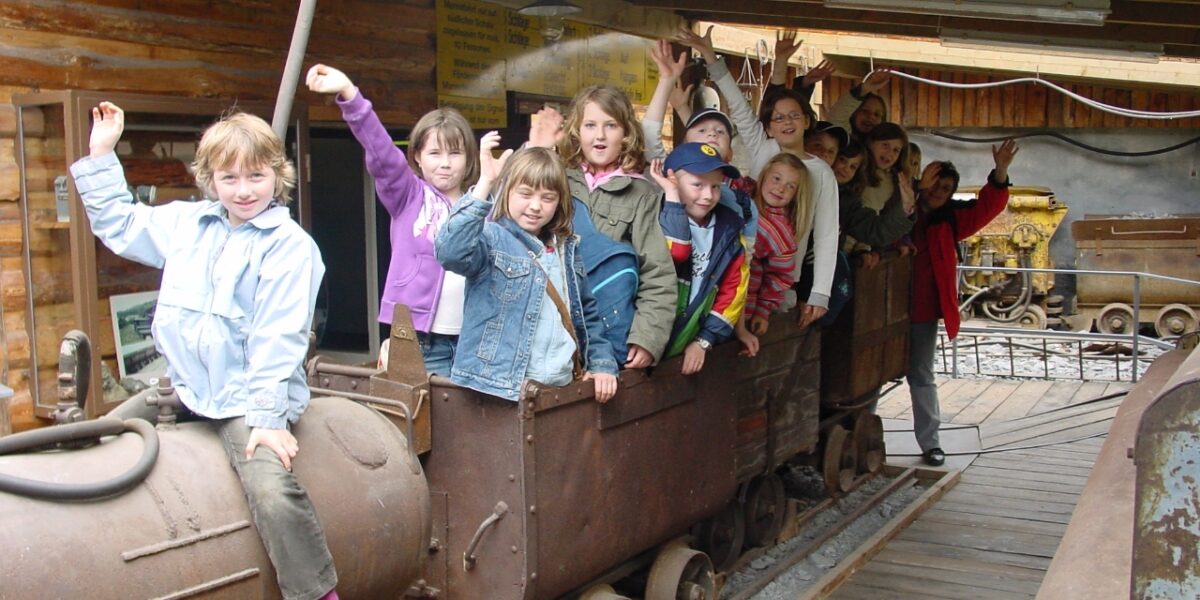 Eine Gruppe von Kindern sitzt und steht auf einem alten Bergwerkswagen in einem Holzgebäude, winkt und lächelt in die Kamera.