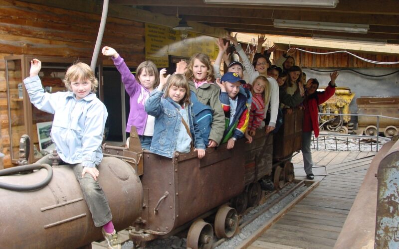 Eine Gruppe von Kindern sitzt und steht auf einem alten Bergwerkswagen in einem Holzgebäude, winkt und lächelt in die Kamera.