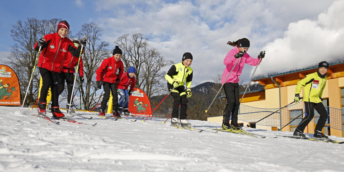 Eine Gruppe von Kindern läuft in farbenfroher Wintersportkleidung auf Skiern auf einem verschneiten Hang, im Hintergrund sind Gebäude und Berge zu sehen.