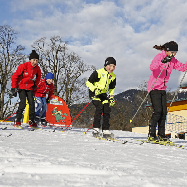 Eine Gruppe von Kindern läuft in farbenfroher Wintersportkleidung auf Skiern auf einem verschneiten Hang, im Hintergrund sind Gebäude und Berge zu sehen.