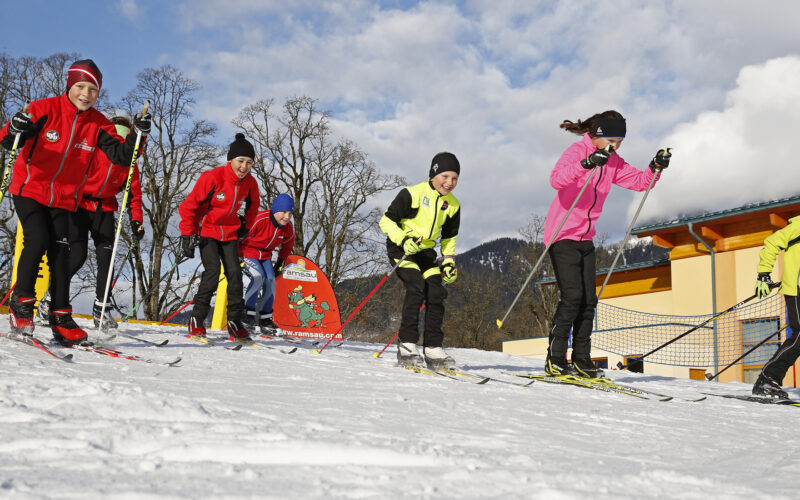Eine Gruppe von Kindern läuft in farbenfroher Wintersportkleidung auf Skiern auf einem verschneiten Hang, im Hintergrund sind Gebäude und Berge zu sehen.