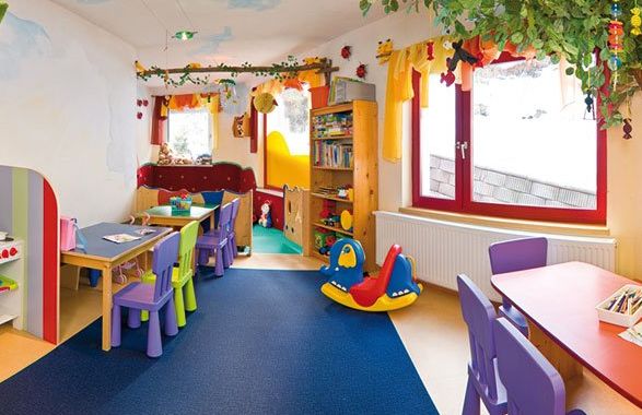 Buntes Kinderspielzimmer mit kleinen Tischen und Stühlen, Spielzeug, Bücherregalen, Spielgeräten und hellem Dekor; natürliches Licht fällt durch die Fenster.