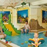 Drei Personen, darunter ein Kind in einem Schwimmer, spielen in einem Kinderbecken mit Wasserspielzeug und einer grünen Rutsche, umgeben von farbenfroher Wasserdekoration.