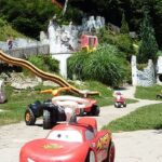Spielplatz im Freien mit Spielzeugfahrzeugen auf einem Weg, Spielgeräten im Stil einer Burg, Rutschen und Grünflächen im Hintergrund.