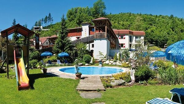 Ein kleiner Außenpool mit Liegestühlen, Sonnenschirmen, einer Spielplatzrutsche und Grünflächen neben einem Hotelgebäude in einer hügeligen Gegend.