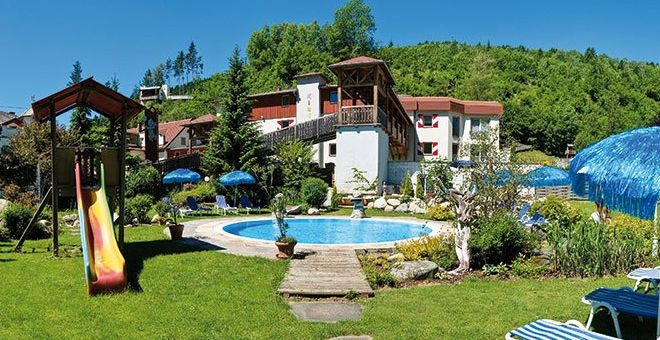 Ein kleiner Außenpool mit Liegestühlen, Sonnenschirmen, einer Spielplatzrutsche und Grünflächen neben einem Hotelgebäude in einer hügeligen Gegend.