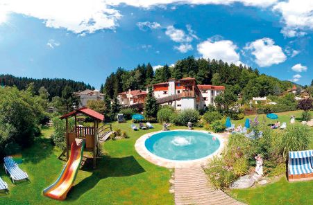 Rundes Freibad und Spielplatz mit Rutsche auf einer Wiese, umgeben von Liegestühlen, Bäumen und Gebäuden unter einem blauen Himmel mit Wolken.