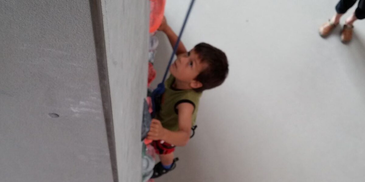 Ein Kind mit einem Klettergurt klettert an einer Indoor-Kletterwand hoch und hält sich dabei an bunten Griffen fest. Im Hintergrund steht eine weitere Person.