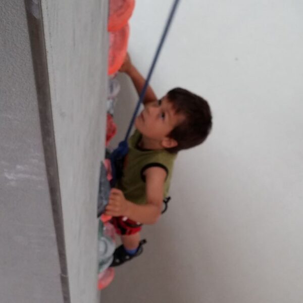 Ein Kind mit einem Klettergurt klettert an einer Indoor-Kletterwand hoch und hält sich dabei an bunten Griffen fest. Im Hintergrund steht eine weitere Person.
