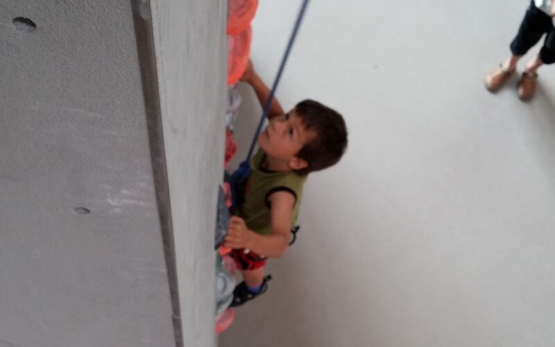 Ein Kind mit einem Klettergurt klettert an einer Indoor-Kletterwand hoch und hält sich dabei an bunten Griffen fest. Im Hintergrund steht eine weitere Person.