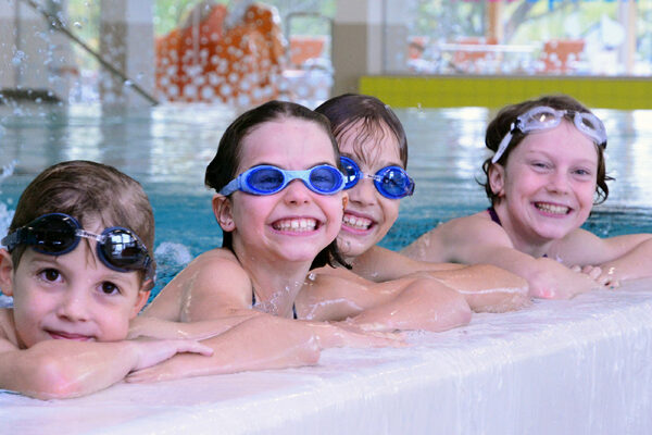 Vier Kinder mit Schwimmbrillen lächeln, während sie sich am Rand eines Hallenbades festhalten. Im Hintergrund plätschert das Wasser.