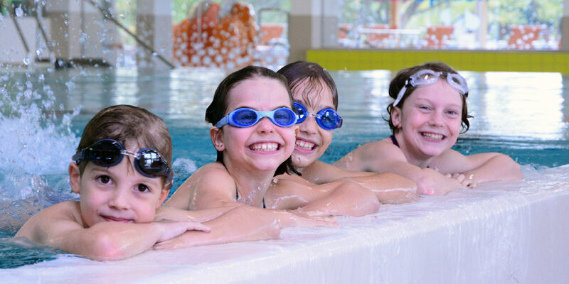 Vier Kinder mit Schwimmbrillen lächeln, während sie sich am Rand eines Hallenbades festhalten. Im Hintergrund plätschert das Wasser.