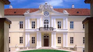 Ein prächtiges, cremefarbenes Gebäude mit rotem Ziegeldach, symmetrischen Fenstern, einem zentralen Balkon und verschnörkelten architektonischen Details unter blauem Himmel.