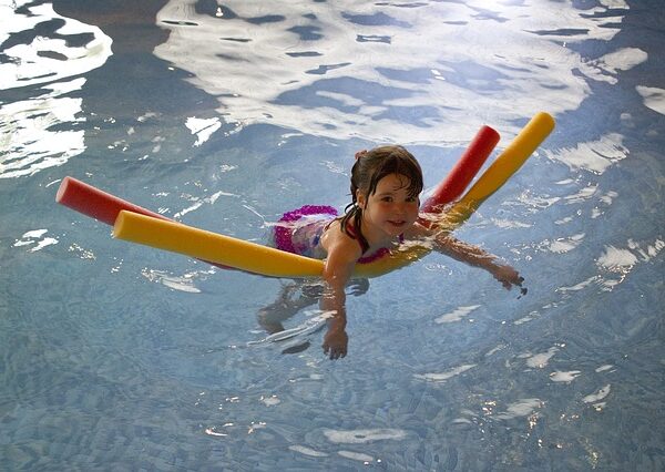 Ein junges Mädchen schwimmt auf zwei bunten Schwimmnudeln in einem Schwimmbecken. Ihre Arme ruhen auf ihnen, und das Wasser plätschert um sie herum.