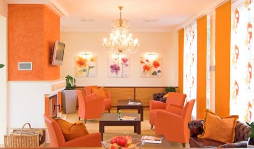 Helle Lounge mit orangefarbenen Stühlen, floraler Wandkunst, einem Kronleuchter und gemusterten Vorhängen. Auch Couchtische und ein Obstkorb sind zu sehen.