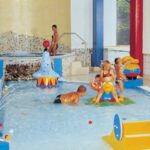 Kinder und Erwachsene spielen und entspannen sich in einem flachen Innenbecken mit buntem Spielzeug und einem kleinen Whirlpool im Hintergrund.
