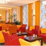 In der hellen Lounge stehen rote und gelbe Sessel, dunkle Holztische und große Fenster mit gemusterten Vorhängen in Orange und Weiß.