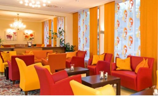 In der hellen Lounge stehen rote und gelbe Sessel, dunkle Holztische und große Fenster mit gemusterten Vorhängen in Orange und Weiß.