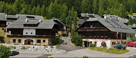 Eine Ansammlung von Gebäuden im alpinen Stil mit dunklen Dächern und Holzbalkonen inmitten grüner Bäume, umgeben von einer gepflasterten Straße und parkenden Autos.
