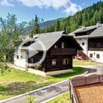 Eine Ansammlung von Häusern im Chalet-Stil in einer bergigen Gegend mit grünen Hügeln, einem Spielplatz mit einer Rutsche im Vordergrund und Blumen in der linken Ecke unter einem teilweise bewölkten Himmel.