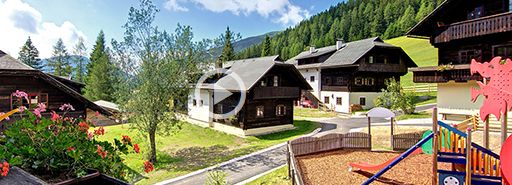 Eine Ansammlung von Häusern im Chalet-Stil in einer bergigen Gegend mit grünen Hügeln, einem Spielplatz mit einer Rutsche im Vordergrund und Blumen in der linken Ecke unter einem teilweise bewölkten Himmel.