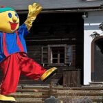 Eine Person in einem bunten Zeichentrickkostüm mit gelbem Gesicht, blauem Hemd, roter Hose und grüner Mütze steht vor einem Holzhaus und hebt einen Arm und ein Bein.