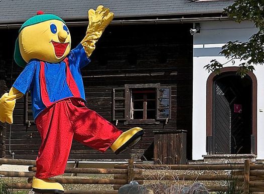 Eine Person in einem bunten Zeichentrickkostüm mit gelbem Gesicht, blauem Hemd, roter Hose und grüner Mütze steht vor einem Holzhaus und hebt einen Arm und ein Bein.