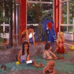 Mehrere Kinder spielen in einem flachen Hallenbad, umgeben von Felsen und tropischen Pflanzen, mit großen Fenstern und roten Wänden im Hintergrund.