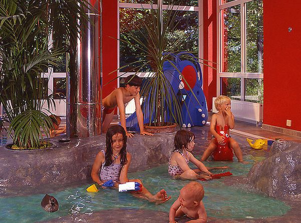 Mehrere Kinder spielen in einem flachen Hallenbad, umgeben von Felsen und tropischen Pflanzen, mit großen Fenstern und roten Wänden im Hintergrund.