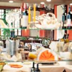 Glasregale mit Flaschen, Gläsern und gefalteten Servietten im Vordergrund; im Hintergrund eines Restaurants sind gedeckte Tische und Pflanzen zu sehen.