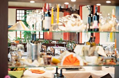 Glasregale mit Flaschen, Gläsern und gefalteten Servietten im Vordergrund; im Hintergrund eines Restaurants sind gedeckte Tische und Pflanzen zu sehen.