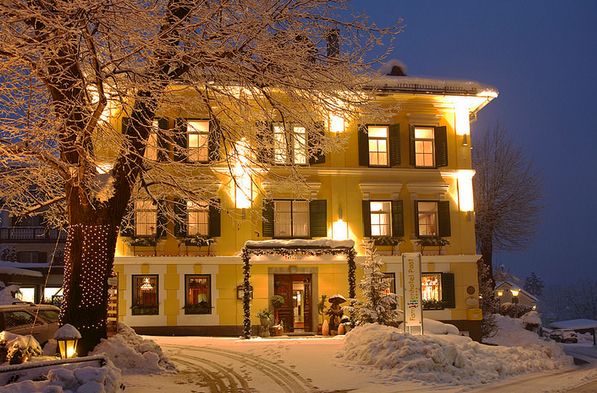 Ein gelbes, dreistöckiges Gebäude mit beleuchteten Fenstern steht an einem Winterabend inmitten schneebedeckter Bäume und Böden.
