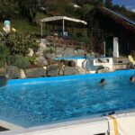 Zwei Personen schwimmen in einem rechteckigen Außenbecken mit einer Wasserrutsche, Felsen und Pflanzen in der Nähe. Im Hintergrund befinden sich Liegestühle und eine Struktur.