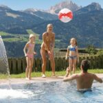 Eine Frau und zwei Mädchen spielen mit einem Strandball in einem Schwimmbad mit einem Mann, mit einer Berglandschaft im Hintergrund.